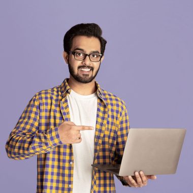 Online work, education, communication concept. Happy Indian guy pointing at laptop over lilac background